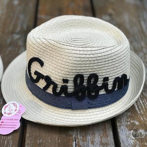Op de afbeelding: Een beige fedora hoed met een donkerblauwe band en de naam "Griffin" in zwart schrift. De hoed is gemaakt van geweven materiaal en ligt op een houten oppervlak. De hoed heeft een ronde kroon en een brede rand.