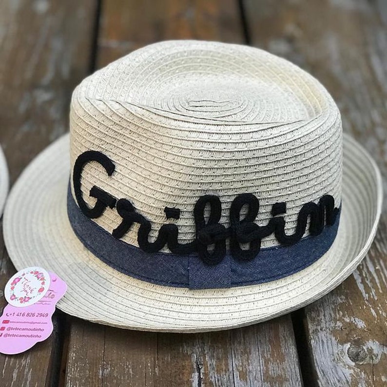 Op de afbeelding: Een beige fedora hoed met een donkerblauwe band en de naam "Griffin" in zwart schrift. De hoed is gemaakt van geweven materiaal en ligt op een houten oppervlak. De hoed heeft een ronde kroon en een brede rand.