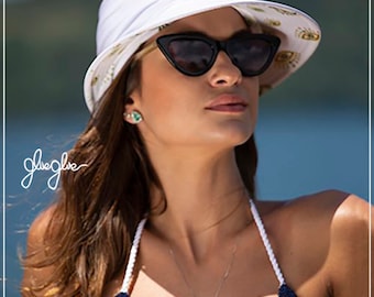 May include: A woman wearing a white sun hat with a gold and white pattern, black sunglasses, and a green heart necklace. The hat has a wide brim and a knotted detail on top. The woman is wearing a blue and white striped bikini top.