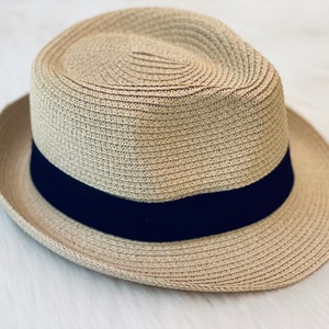 Op de afbeelding: Een lichtbruine fedora hoed met een donkerblauwe band. De hoed is gemaakt van geweven stro en heeft een klassieke, gestructureerde vorm. De rand is ongeveer 5 cm breed.