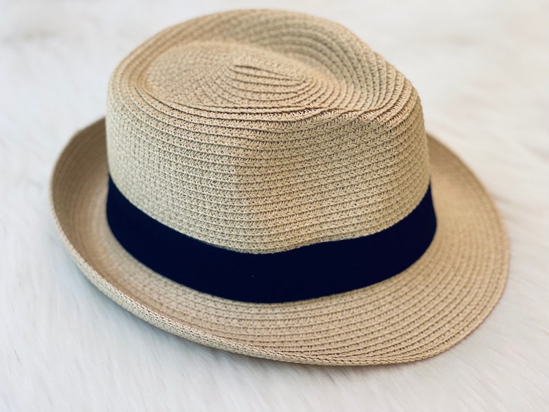 Op de afbeelding: Een lichtbruine fedora hoed met een donkerblauwe band. De hoed is gemaakt van geweven stro en heeft een klassieke, gestructureerde vorm. De rand is ongeveer 5 cm breed.