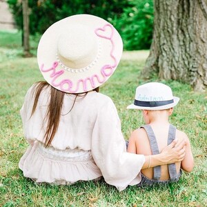 Op de afbeelding: Een vrouw en een kind zitten op het gras. De vrouw draagt een strohoed met brede rand met het woord "Mama" in roze script. Het kind draagt een witte hoed met de naam "Adrian" in blauw script.