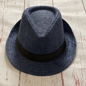 Op de afbeelding: Een marineblauwe fedora hoed met een zwarte band. De hoed is gemaakt van geweven materiaal en heeft een klassieke vorm. De hoed ligt op een lichtgekleurd houten oppervlak.