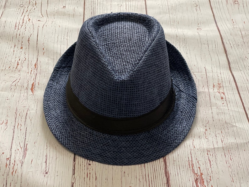 Op de afbeelding: Een marineblauwe fedora hoed met een zwarte band. De hoed is gemaakt van geweven materiaal en heeft een klassieke vorm. De hoed ligt op een lichtgekleurd houten oppervlak.