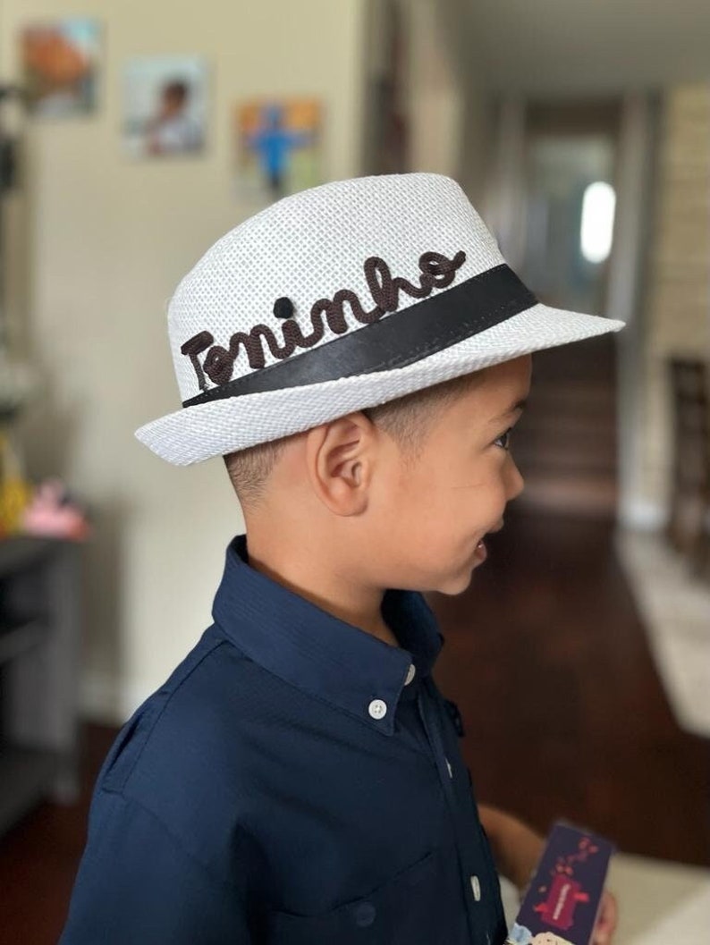 Op de afbeelding: Witte fedora hoed met een zwarte band en de naam "Toninho" in bruin schrift. De hoed is gemaakt van een geweven materiaal en heeft een klassieke fedora vorm. De hoed wordt gedragen door een persoon die een marineblauw overhemd draagt.