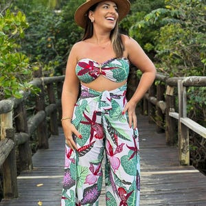 Könnte beinhalten: Eine Frau trägt einen weißen und grünen Tropenprint-Jumpsuit mit einem roten floralen Bandeau-Top. Sie trägt einen breitkrempigen Strohhut und rote Sandalen. Sie steht auf einem Holzsteg.