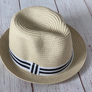Op de afbeelding: Een beige fedora hoed met een zwart-wit gestreepte band. De hoed is gemaakt van geweven materiaal en heeft een klassieke fedora vorm. De hoed ligt op een lichtgekleurd houten oppervlak.