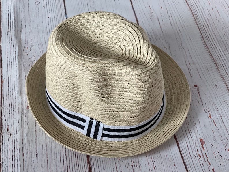 Op de afbeelding: Een beige fedora hoed met een zwart-wit gestreepte band. De hoed is gemaakt van geweven materiaal en heeft een klassieke fedora vorm. De hoed ligt op een lichtgekleurd houten oppervlak.