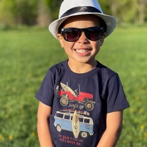 Op de afbeelding: Een kind draagt een witte fedora hoed met een zwarte band en een zwarte zonnebril en glimlacht in de camera. Het kind draagt een marineblauw t-shirt met een rode jeep en een blauwe busje. De tekst op het shirt luidt: "De zee roept en ik moet gaan."