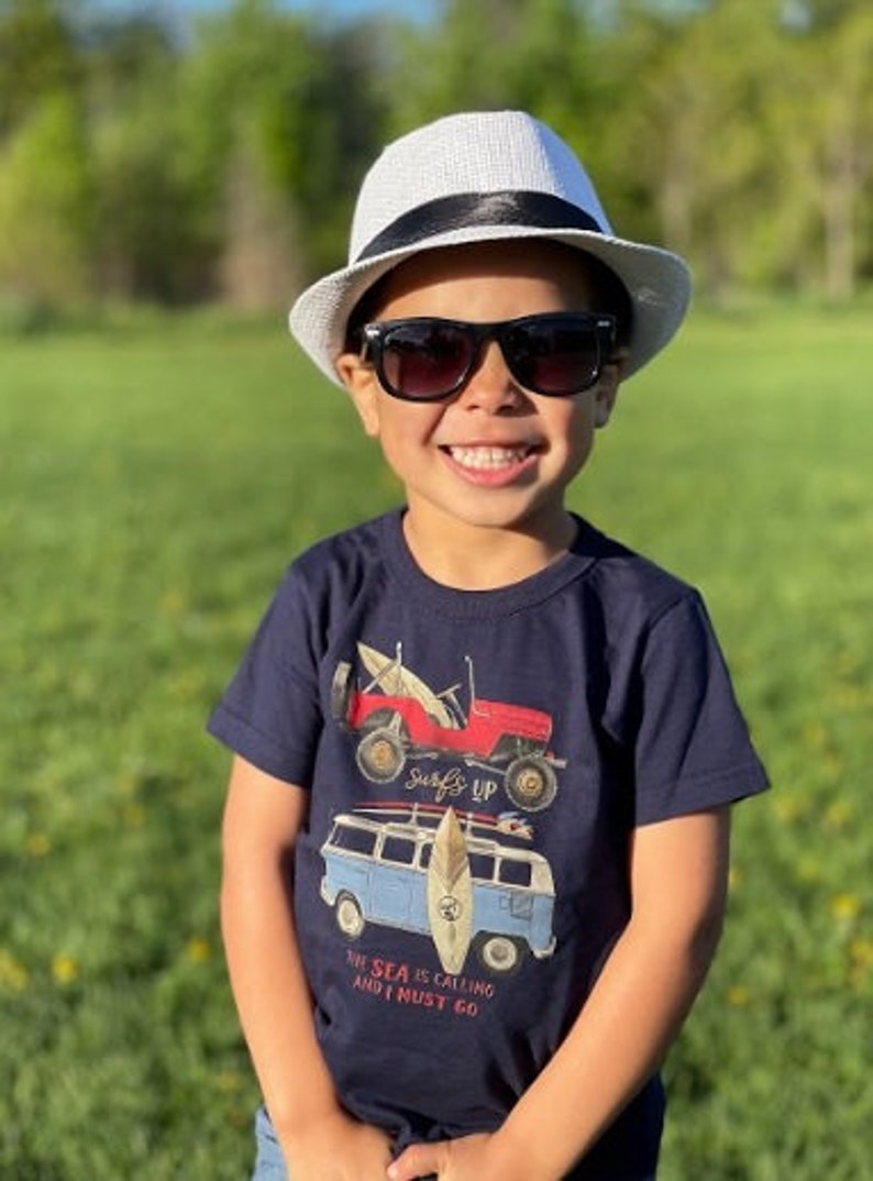 Op de afbeelding: Een kind draagt een witte fedora hoed met een zwarte band en een zwarte zonnebril en glimlacht in de camera. Het kind draagt een marineblauw t-shirt met een rode jeep en een blauwe busje. De tekst op het shirt luidt: "De zee roept en ik moet gaan."