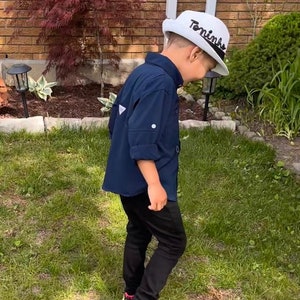 Op de afbeelding: Een persoon draagt een marineblauw overhemd, een zwarte broek en kleurrijke sneakers. De persoon draagt ook een witte hoed met de naam "Toninho" erop. De sc&egrave;ne speelt zich buiten af, met groen gras en een bakstenen muur.