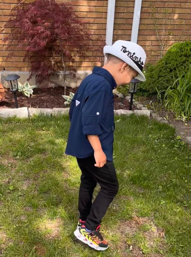 Op de afbeelding: Een persoon draagt een marineblauw overhemd, een zwarte broek en kleurrijke sneakers. De persoon draagt ook een witte hoed met de naam "Toninho" erop. De sc&egrave;ne speelt zich buiten af, met groen gras en een bakstenen muur.