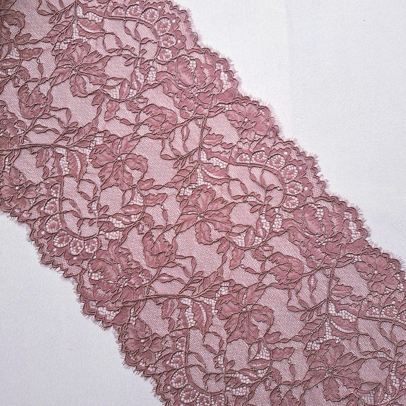 May include: Dusty rose lace fabric with a floral pattern. The lace features detailed flower and leaf designs, with a scalloped edge. The fabric is a soft, muted pink color, suitable for various craft and sewing projects.