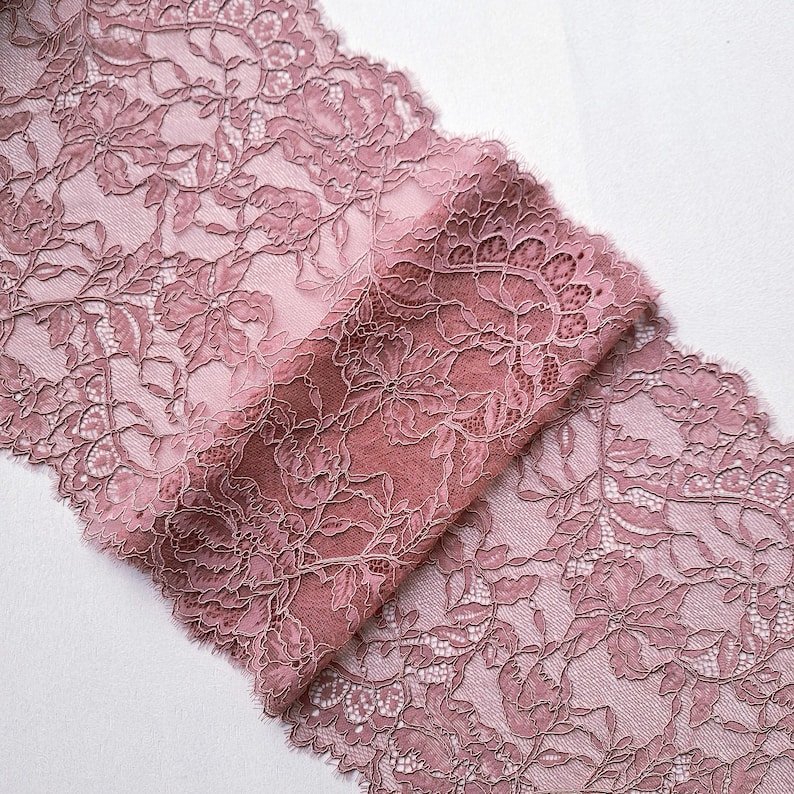 May include: Dusty rose lace fabric with a floral pattern. The delicate lace is folded over, revealing the intricate design and texture. The fabric is ideal for sewing, crafting, or adding a touch of elegance to any project.