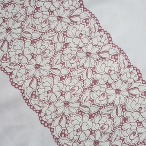 May include: A close-up of a pink lace trim with a white floral pattern. The lace is delicate and intricate, with a scalloped edge.