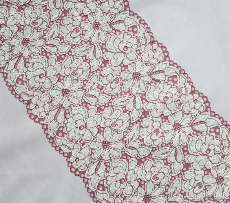 May include: A close-up of a pink lace trim with a white floral pattern. The lace is delicate and intricate, with a scalloped edge.