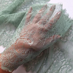 May include: A close-up of sage green lace fabric. The fabric is held up, showing the intricate floral pattern and detailed edge. The light passing through highlights the delicate texture and design of the lace, creating a translucent effect.