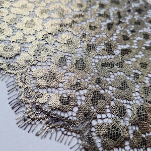 Può includere: Primo piano di un tessuto di pizzo oro e nero. Il pizzo presenta un motivo floreale con una lucentezza metallica. Il design intricato è visibile, con una trama delicata e aperta. Il bordo del tessuto ha un bordo smerlato.