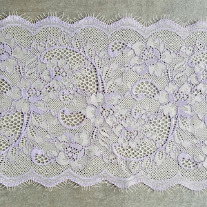 Op de afbeelding: Een close-up van een delicate lavendelkleurige kantversiering met een bloemenpatroon. De kant heeft een gegolfde rand en is gemaakt van een fijn, lichtgewicht materiaal.