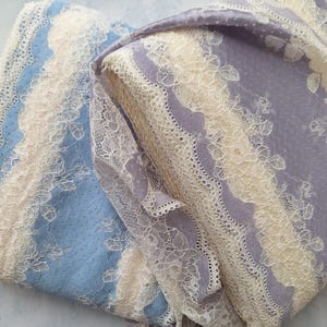 May include: Close-up of two fabric pieces with a lace overlay. One piece is light blue, the other is lavender. Both have a cream-colored lace border. The lace features a floral design. The fabrics are folded, showing the texture and pattern.