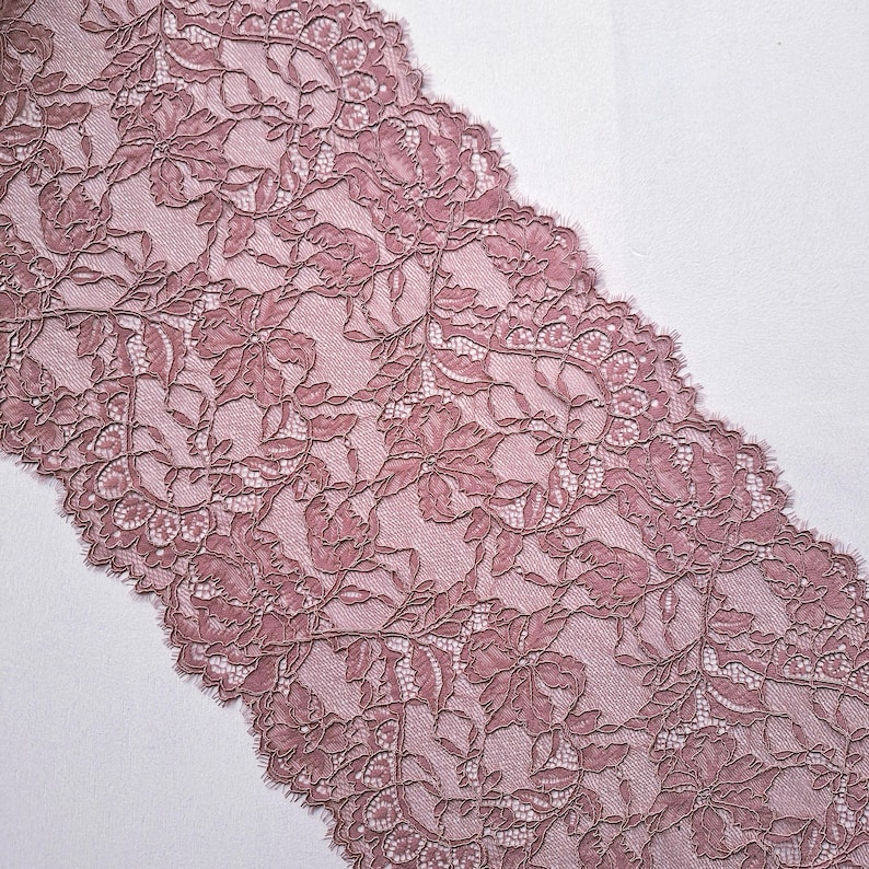 May include: Dusty rose lace fabric with a floral pattern. The lace features detailed designs of flowers and leaves, with a scalloped edge. The fabric is likely used for sewing, crafts, or apparel.