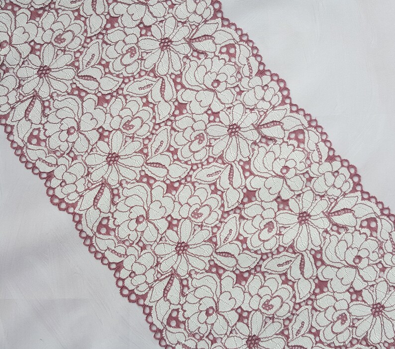 May include: A close-up of a pink lace trim with a white floral pattern. The lace is delicate and intricate, with a scalloped edge.