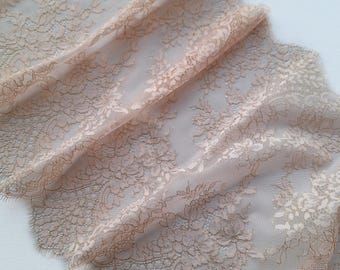 May include: A delicate, sheer lace fabric in a soft peach color. The lace features a floral pattern and a scalloped edge. The fabric is draped, showing its texture and intricate design, suitable for apparel or crafts.