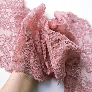 May include: Close-up of a delicate, dusty rose lace fabric. The lace features a floral pattern with intricate details and a scalloped edge. The fabric is soft and flowing, ideal for apparel or craft projects.