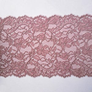 May include: Close-up of a dusty rose lace fabric. The lace features a floral pattern with detailed leaves and flowers. The edges are scalloped, adding a delicate touch. The fabric is likely used for apparel or craft projects.