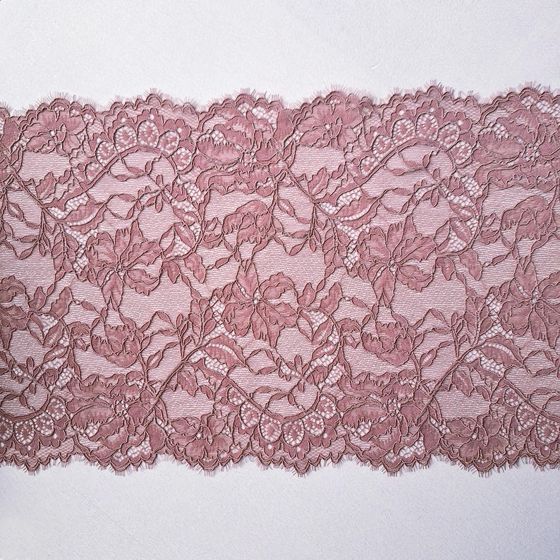 May include: Close-up of a dusty rose lace fabric. The lace features a floral pattern with detailed leaves and flowers. The edges are scalloped, adding a delicate touch. The fabric is likely used for apparel or craft projects.