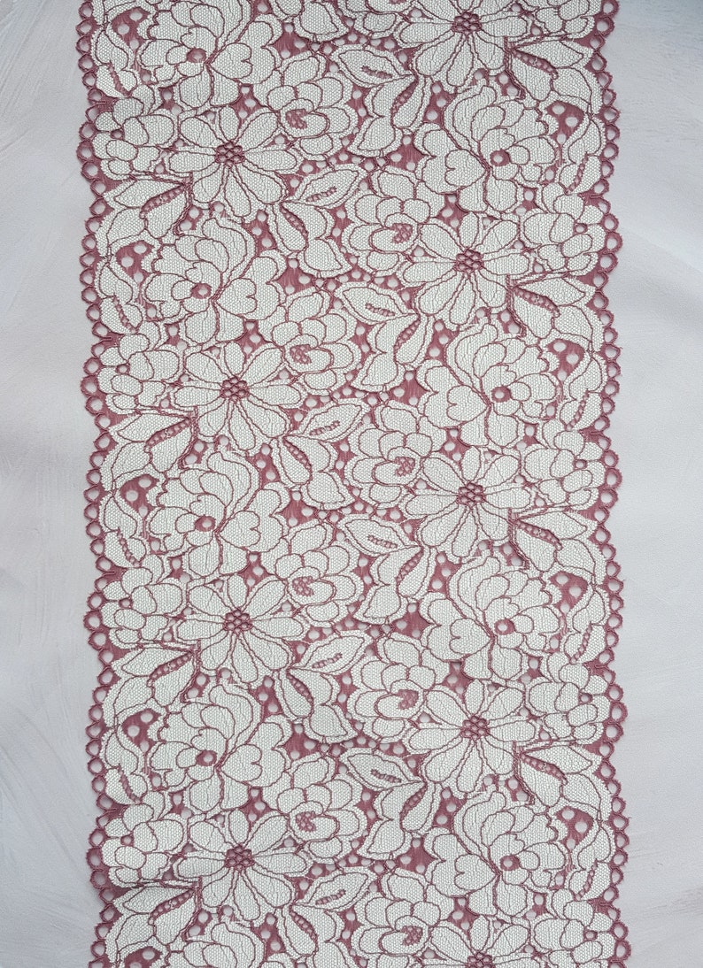 May include: A close-up of a pink lace trim with a white floral pattern. The lace is delicate and intricate, with a scalloped edge.