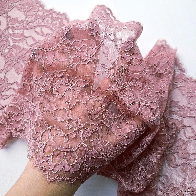 May include: Close-up of a delicate, dusty rose lace fabric. The lace features a floral pattern with intricate details. The fabric is sheer, allowing light to pass through, and has a scalloped edge. The overall effect is soft and romantic.