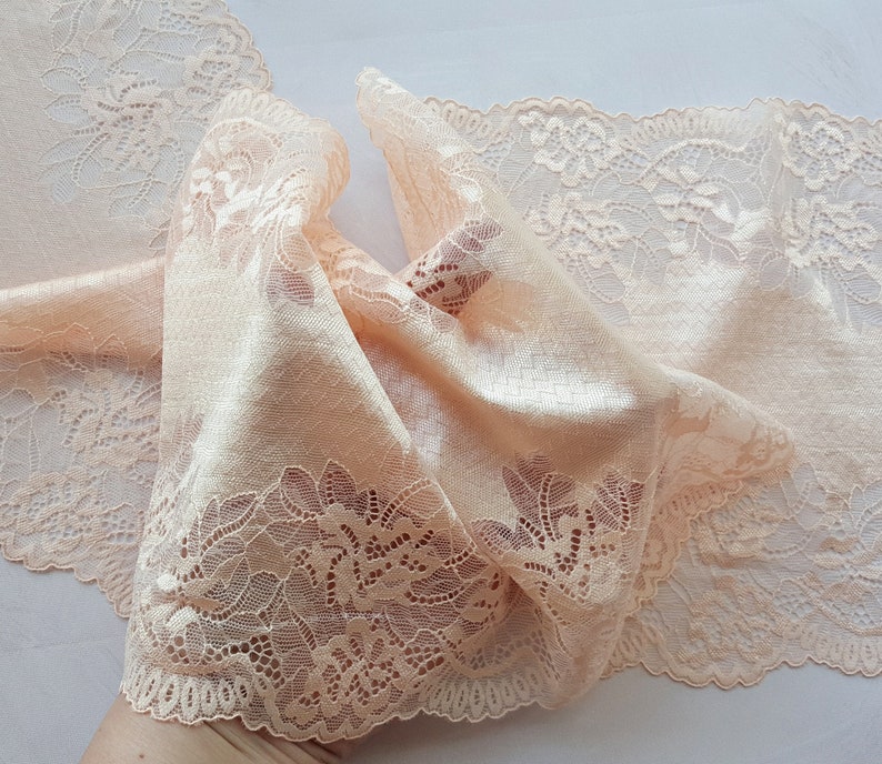 May include: Peach colored lace trim with a delicate floral pattern. The lace is soft and flowing, with a scalloped edge.