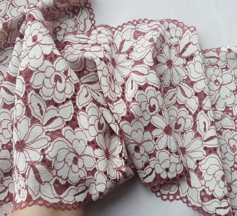May include: A close-up of a pink and white floral lace trim. The lace has a delicate floral pattern and a scalloped edge.