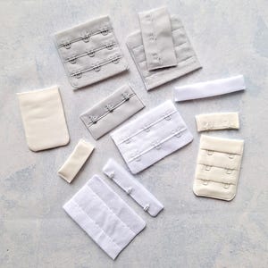 Pode incluir: Conjunto de extensões de alça de sutiã e fechos de gancho e olho em branco, bege e cinza. As extensões são de elástico e têm ganchos de metal. Os fechos de gancho e olho são de plástico e têm ganchos de metal.