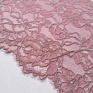 May include: Close-up of a dusty rose lace fabric with a floral pattern. The delicate lace features intricate details and a scalloped edge. The fabric is soft and textured, perfect for crafting or adding a touch of elegance to any project.