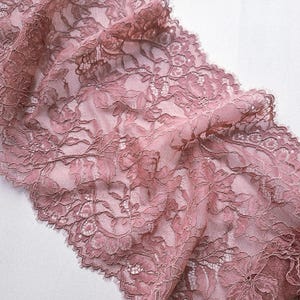 May include: Close-up of a dusty rose lace fabric with a floral pattern. The delicate lace features intricate details and a scalloped edge. The fabric is semi-sheer, allowing the background to show through, creating a soft, romantic aesthetic.