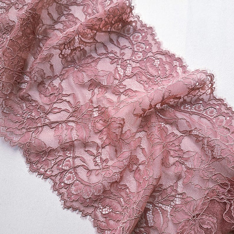 May include: Close-up of a dusty rose lace fabric with a floral pattern. The delicate lace features intricate details and a scalloped edge. The fabric is semi-sheer, allowing the background to show through, creating a soft, romantic aesthetic.