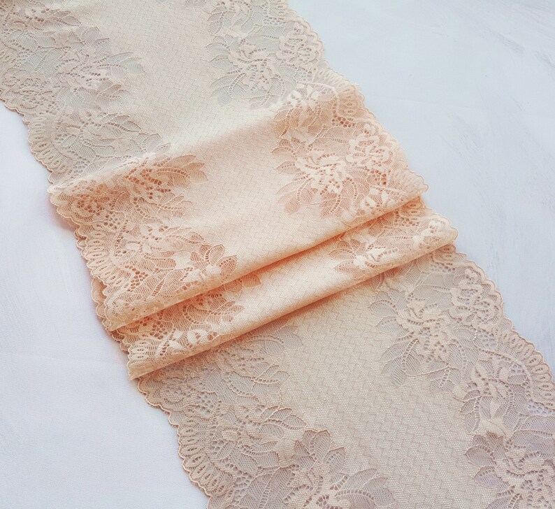 May include: Peach colored lace trim with a floral pattern. The lace is folded in thirds and is on a white background.