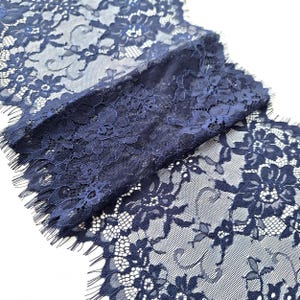 Puede incluir: Primer plano de tela de encaje azul marino con un patrón floral. El delicado encaje presenta diseños intrincados y un borde con flecos. La tela está doblada, mostrando la textura y los detalles del encaje. Ideal para proyectos de costura, manualidades o fines decorativos.