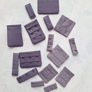 Könnte beinhalten: Ein Set aus lilafarbenen BH-Verlängerungen und Haken- und Ösenverschlüssen. Die Verlängerungen bestehen aus einem weichen, dehnbaren Stoff und sind in verschiedenen Längen erhältlich. Die Haken- und Ösenverschlüsse sind aus Metall und in verschiedenen Größen erhältlich.