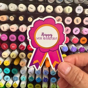 May include: A die-cut sticker shaped like a rosette ribbon with a white center, a bright orange inner border, and a magenta outer edge. The text "Happy WORK ANNIVERSARY!" is printed in purple in the center of the sticker, which features two downward-pointing ribbon tails with orange and magenta coloring.