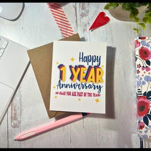 May include: A white greeting card with a brown envelope featuring the text Happy 1 YEAR Anniversary SO GLAD YOU ARE PART OF THE TEAM! in blue, yellow, and red lettering with small yellow star accents on a light wood surface.