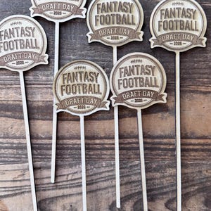 Puede incluir: Cinco adornos para pastel de madera clara, cada uno con un diseño en forma de escudo con un icono de balón de fútbol grabado, el texto FANTASY FOOTBALL DRAFT DAY y el año 2026. Cada adorno está unido a un palo de madera largo y fino.