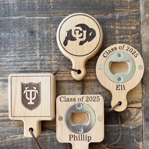 May include: Three wooden bottle openers with a brown leather cord. The top opener is round with a University of Wyoming logo and the text "Class of 2025". The middle opener is square with a University of Pennsylvania logo. The bottom opener is square with a bottle opener, the text "Class of 2025", and the name "Phillip".