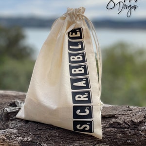 May include: A beige drawstring bag with the word "SCRABBLE" printed vertically in black on the side. The bag is made of a natural fabric and has a drawstring closure. The background is blurred, showing a body of water and trees.