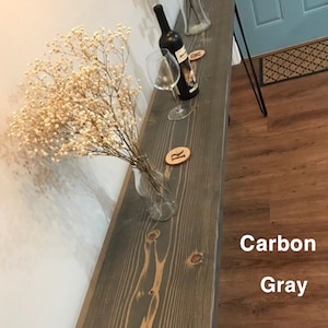 May include: A long, narrow wooden console table with black metal hairpin legs. The table top is stained a dark gray color with a wood grain pattern. The table is decorated with a vase of dried flowers and a wine bottle and glass.