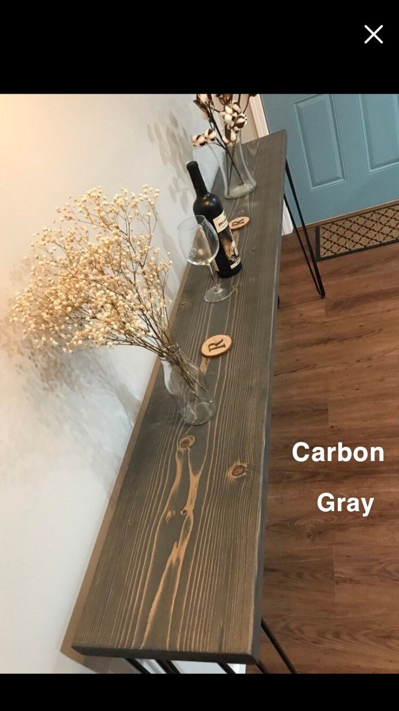 May include: A long, narrow wooden console table with black metal hairpin legs. The table top is stained a dark gray color with a wood grain pattern. The table is decorated with a vase of dried flowers and a wine bottle and glass.