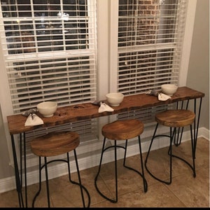 May include: A long wooden table with a live edge top and black hairpin legs. Three black metal bar stools with wooden seats are placed in front of the table. Two white bowls are on the table.