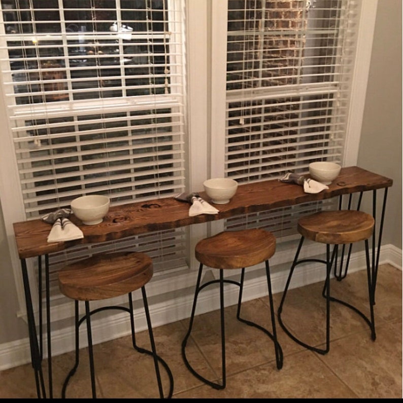 May include: A long wooden table with a live edge top and black hairpin legs. Three black metal bar stools with wooden seats are placed in front of the table. Two white bowls are on the table.